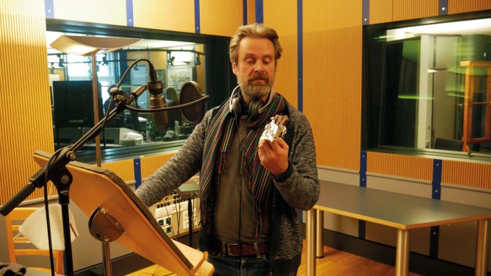 Matthias Matschke schaut auf Schokolade, im Studio bei der Aufnahme Mitmachhörspiel zur ARD-Kinderradionacht 2020 (Quelle: rbb/OHRENBÄR/Birgit Patzelt)