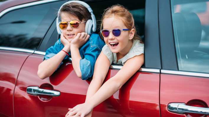 Ein Junge mit Kopfhörer und ein Mädchen, beide mit einer Sonnebrille, schauen aus einem geöffneten Autofenster (Quelle: imago/Pond 5)
