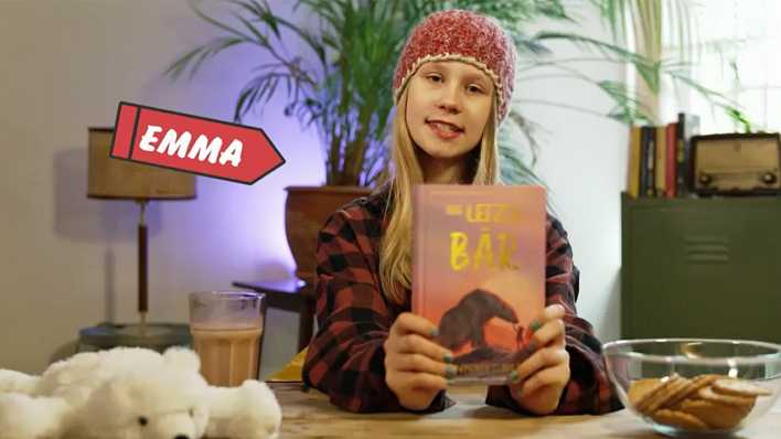 Buchtesterin Emma zeigt das Buch "Der letzte Bär", sitzt am Schreibtisch, darauf Kekse und ein weißer Stoffbär (Quelle: KiKa)