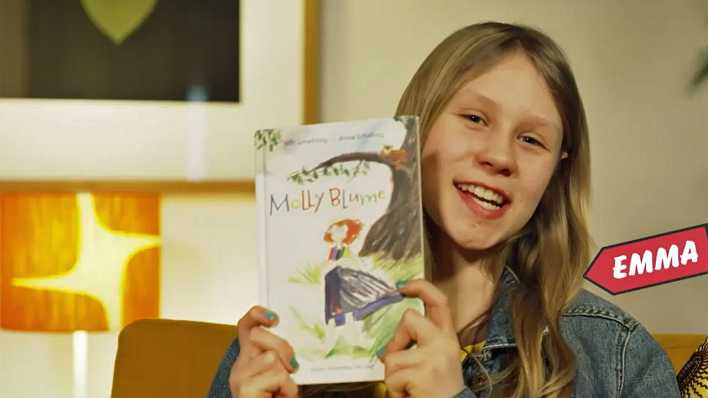 Buchtesterin Emma zeigt das Buch "Molly Blume", vor einer hellen Wand, davor Stehlampe und Bilderrahmen (Quelle: KiKa)